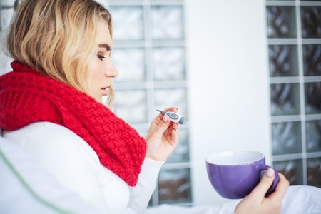 High temperature. Young woman looking at thermometer while holding it in hands and lying in bed