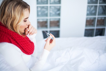 High temperature. Young woman looking at thermometer while holding it in hands and lying in bedの写真素材