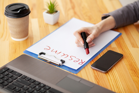 Job search. Female hand writes resume with lipstick on white sheet of paper. Wooden office desk with laptop, smartphone and suppliesの写真素材