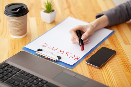 Job search. Female hand writes resume with lipstick on white sheet of paper. Wooden office desk with laptop, smartphone and supplies