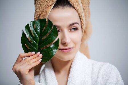 Beautiful woman in a white coat with a towel on her head and clear skin holds a green leaf
