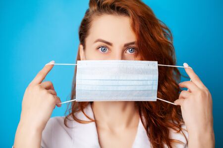 Woman holding mask to cover mouth and nose to protect against virus on blue background.の写真素材