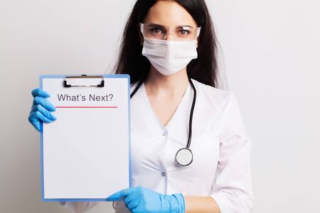 Doctor in a gloves, mask and glasses holds sheet with text whats next.の写真素材
