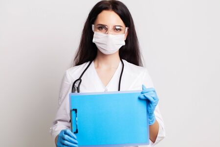 Young female doctor in a mask holds a blank sheet of paper with space for textの写真素材