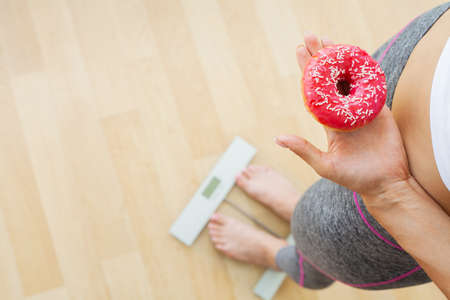 Diet concept, woman measures weight on electronic scales while holding calorie donut