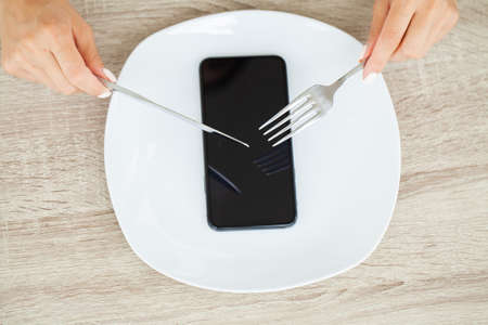 Dependence on the phone, close up a womans hands holding a fork and a knife over the phone on plateの写真素材