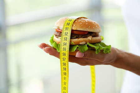 Diet concept, woman holding harmful fat burger with yellow measuring tapeの写真素材