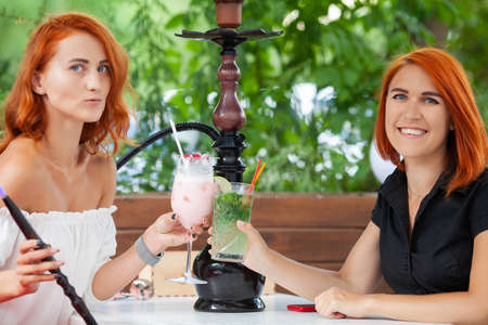 Two young women smoke a hookah and enjoy cocktails in cafe on the street