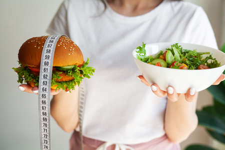 Beautiful young woman decides eating hamburger or fresh salad in kitchen.の写真素材
