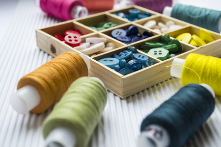 Various colored threads with clothes buttons in wooden box on white cardboard background. Closeup photo. Macro shotの写真素材