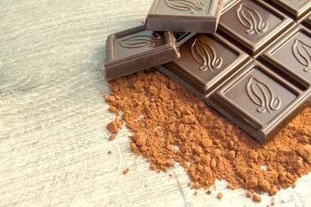 bar of chocolate on cocoa powder. pieces of chocolate lie next to the bar. powder scattered on wood