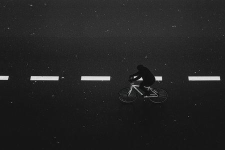 A lone cyclist glides silently along wet pavement, surrounded by reflective white lines under soft streetlights at night.の素材