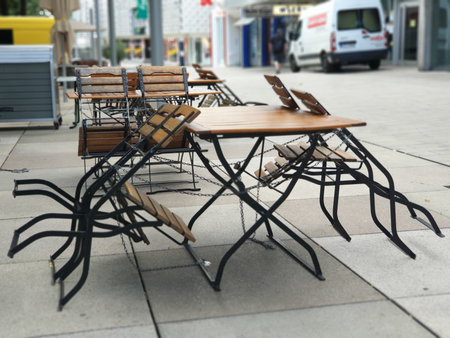 The photo shows wooden tables and chairs stacked and chained on a city street. In the background, there are buildings and a parked car.の写真素材
