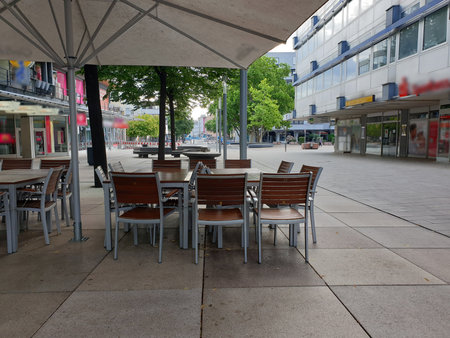 A cozy street café with wooden tables under a large umbrella. The pedestrian zone is framed by modern buildings and greenery. A relaxed and inviting atmosphere.の写真素材