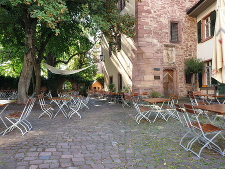 The photo shows a cozy café with wooden furniture outdoors. Surrounded by green trees and historic stone buildings, it exudes a calm and relaxing ambiance.の写真素材
