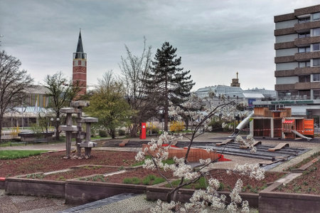 An urban scene featuring a tower surrounded by trees. In the center is a modern sculpture, and to the right, a playground with a slide and play structures.の写真素材