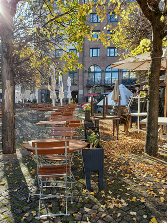 A cozy outdoor cafÃ© is surrounded by trees with golden leaves. A cobblestone path, tables, and gentle shadows create a warm autumn atmosphere.の写真素材