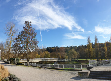 Autumn park with a bridge over a river. Trees with fallen leaves, clear sky with clouds. Path, benches, and forest in the background.の写真素材
