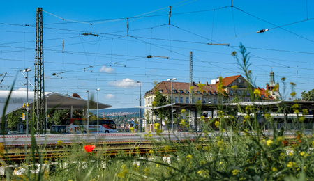 Pforzheim Germany Mai 31, 2018  Sunlit Train Station Platforms And Tracks, Wildflowers Foreground, Historic Station Building, Distant Mountains, Web Of Overhead Wires,のeditorial素材