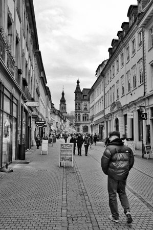 Heidelberg Germany January 24, 2026 Solitary Walker In Puffer Jacket Walking Down Narrow Street Toward Distant Spire, Cobblestone Perspective And Lined Facades Commuter Mood With Cold Light, Heidelberg Germany January ,のeditorial素材