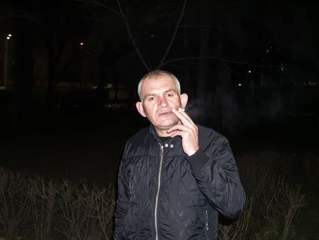 A man with gray hair smokes a cigarette at night on the street. People background image. Father's...