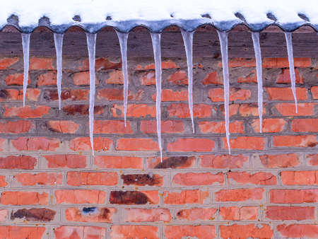 Icicles of frozen water on the roof of a house with a brick wall. Snow melting. Winter. Spring drops. Ice icicle. Frozen water. Brick wall. Frosty frost. Winter season. Roof of the house.の写真素材