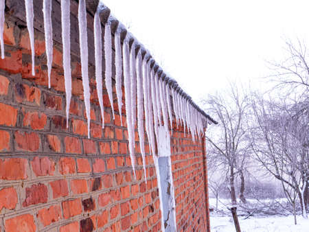 Icicles of frozen water on the roof of a house with a brick wall. Snow melting. Winter. Spring drops. Ice icicle. Frozen water. Brick wall. Frosty frost. Winter season. Roof of the house.の写真素材