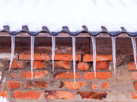 Icicles of frozen water on the roof of a house with a brick wall. Snow melting. Winter. Spring drops. Ice icicle. Frozen water. Brick wall. Frosty frost. Winter season. Roof of the house.の写真素材