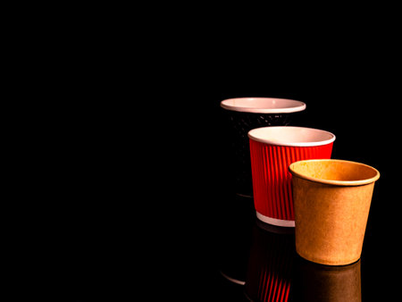 Disposable cardboard coffee cups on a black background. Cardboard colored cups. Disposable tableware. Coffee drinks. Paper dishes for drinks. Hot tea. Black background. Darkness.の写真素材