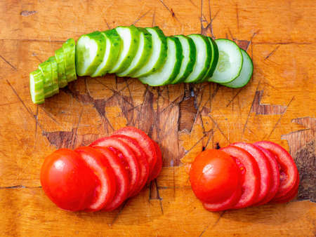 Sliced cucumber and tomato on a wooden cutting board. Cooking vegetables. Green cucumber. Red tomato. Vegetable food. Wooden cutting board. Vegetarian food. Home kitchen. Fresh vitamins.の写真素材