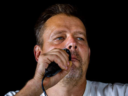 A middle-aged man cuts his gray beard with a shaving trimmer. Mustache and beard clipper. Adult. Male hairdresser. Man's face. Aged people. Hairdresser's tool. Facial care.の写真素材