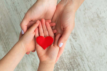 Red heart in child and mother hands on wooden background. Health care and family concept.の写真素材
