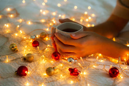 Womans hands holding a cup of tea in bed. Christmas lights on, cozy home atmosphere.