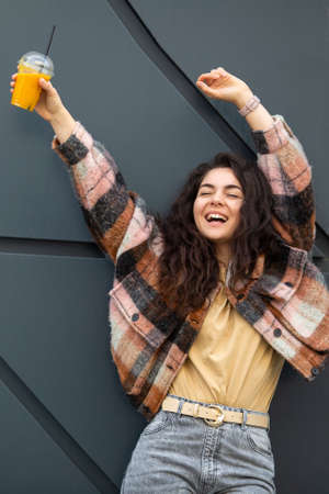 Young stylish woman with juice cup standing over gray background. Portrait of a smiling woman with...