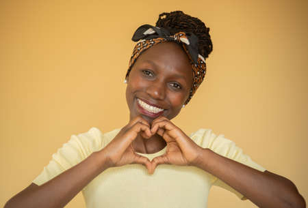 Young woman making a heart shaped symbol while smiling with a yellow backgroundの写真素材