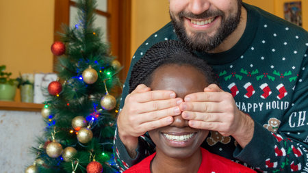 Happy man covering eyes of young african woman with his hands happy receiving a christmas gift in front of the christmas treeの写真素材
