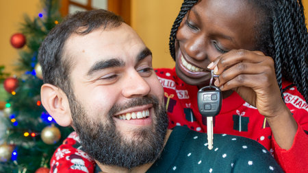 Interracial couple enjoying a key car as a christmas present wearing christmas sweaters in front of...