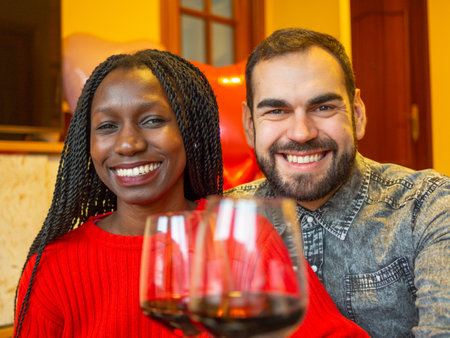 portrait of an interracial couple looking at camera laughing with glasses of red wine enjoying valentines dayの写真素材