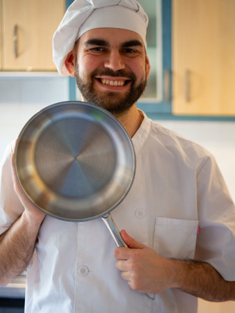 Portrait of a young bearded chef smiling while pointing a stainless pan looking at cameraの写真素材