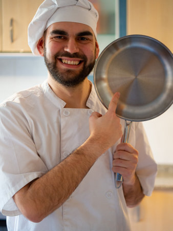 Portrait of a young bearded chef smiling while pointing a stainless pan looking at cameraの写真素材