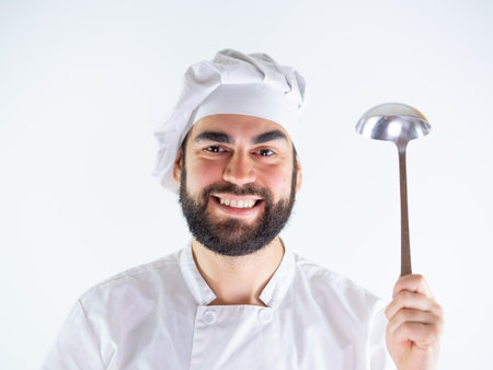 Young male chef showing a metal ladle while smiling and looking at camera. Isolated on a white backgroundの写真素材