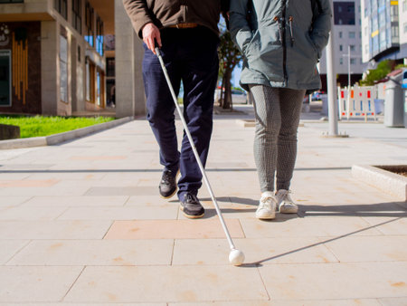 Blind man walking on the pavement using his cane and a woman providing guidance.