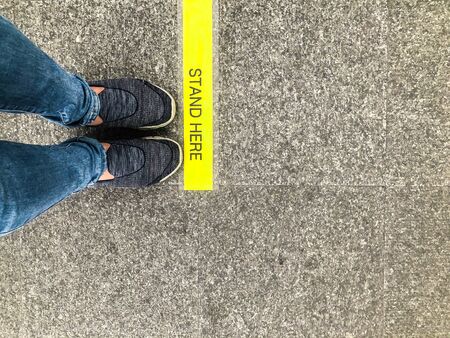Feet and shoes of person standing in front of the yellow sign âstand hereâ on the floor waiting for a queue for keeping distance away from other people during Covid-19 pandemic outbreakの写真素材
