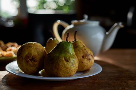 Appetizing pear pham with drops of water on them. The white teapot and plate with vegetable snack on a background in dark key. The sun is shining to the window.の写真素材