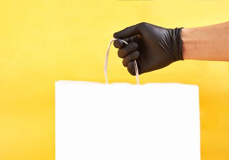 Man's hand holds white paper package on a yellow background. Space for text. Bag with handles