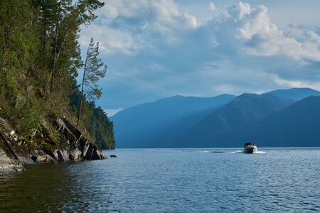 The boat is sailing at mountains lake. Rocks with trees in the left corner. Altai mountains, Teletskoye lakeの写真素材