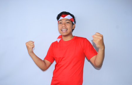 portrait of excited indonesian young man raised his fist to celebrates his succeed or goal in indonesia independence day wearing red t shirt and headband. young man show yes gesture for his achievement and satisfactionの写真素材