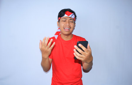 sad indonesian young man is crying when looking to his smartphone, wearing red t shirt and headband isolated in whiteの写真素材