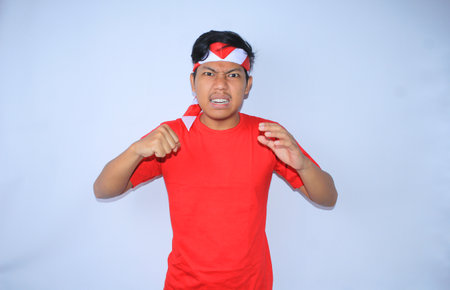 angry indonesian young man raising hands to hit his palm with his fist wearing red t shirt and headband in indonesia independence dayの写真素材