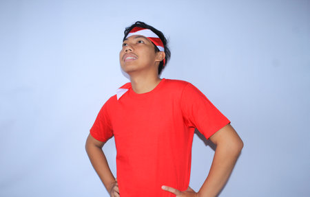 indonesian young man is smiling with hands on waist wearing red t shirt and headband to celebrates indonesia independence day, looking awayの写真素材
